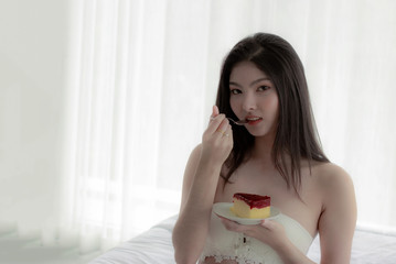 Beautiful Asian woman sit and relax in the bedroom. Happily having a snack on the bed. In the hands beautiful girl have a cake. Woman wearing white bra. Sitting and eating cake with deliciousness.