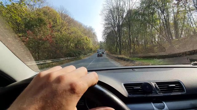Male hands on the steering wheel of the car. View the driver's eyes on the road. Travel by car on the highway between the autumn trees.