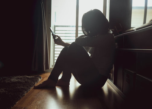 Asian woman sitting on wood floor beside window light in a dark room. Hold and bend head to look at mobile phone screen to check message, chat bad news or wait for a reply by feeling sad or hopeless