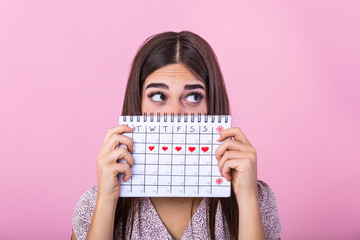 Portrait of a funny young girl in hiding behind a menstrual periods calendar and looking away at copy space isolated over pink background. Female Period calendar - obrazy, fototapety, plakaty