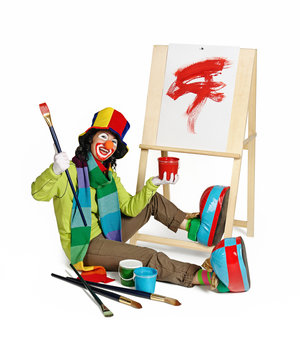 Female clown with big brush in her hands, sitting by  easel on floor. Fun face, makeup, hat and big shoes. Studio shot.