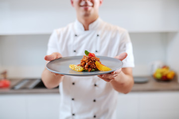 Close up of Caucasian chef in uniform holding plate with salmon and orange fruit. Selective focus on plate.