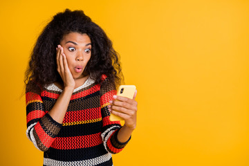 Portrait of astonished impressed afro american girl use her cellphone read feedback feed news on social network copyspace stare stupor wear striped jumper isolated over yellow color background