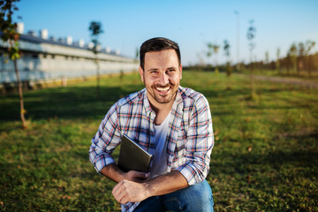 Naklejka na ściany i meble Handsome smiling caucasian farmer in plaid shirt and jeans crouching outdoors with tablet in hands. In background are barn and trees.