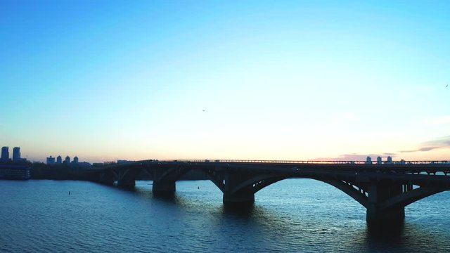 Traffic on the Metro bridge in Kyiv from the left bank to the right. The movement of trains and cars on the bridge over the Dnipro River in the early morning at dawn.