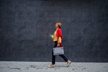 Side view of charming caucasian fashionable senior woman carrying bag and groceries while passing by gray wall.