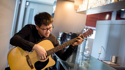 Young Asian man guitarist playing acoustic guitar at bar counter in the cafe. Practicing string...