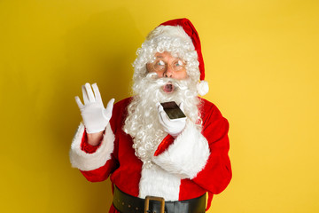 Santa Claus with modern gadgets isolated on yellow studio background. Caucasian male model in traditional costume. Concept of Christmas, New Year's, winter mood, technologies. Records voice messege.