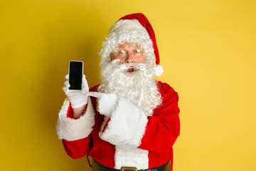 Santa Claus with modern gadgets isolated on yellow studio background. Caucasian male model in traditional costume. Concept of Christmas, New Year's, winter mood, technologies. Shows empty phone screen