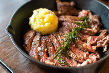 Cast iron plate with sliced grilled rib-eye cooking a medium-rare steak with mashed potato and sauce in saucer over table.