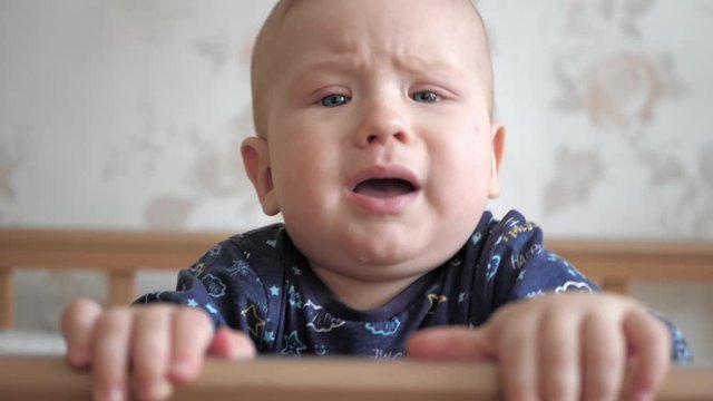 Сute baby with blue eyes stands and screams, freaks out, cries in wooden crib. The child is dressed in blue shirt and holds on to the crib. Slow motion view