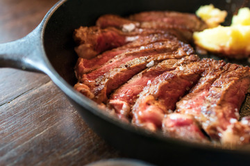 Cast iron plate with sliced grilled rib-eye cooking a medium-rare steak with mashed potato and sauce in saucer over table.
