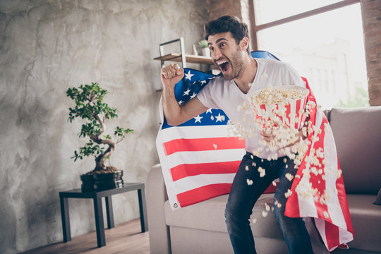 Photo of multiethnic crazy arab guy sitting sofa hold popcorn bucket raising fists supporting usa football team match american flag on shoulders coat flat room indoors