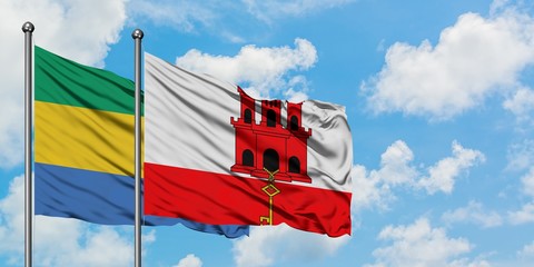 Gabon and Gibraltar flag waving in the wind against white cloudy blue sky together. Diplomacy...
