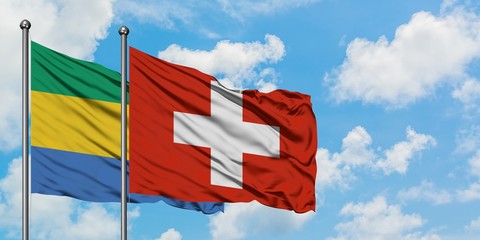 Gabon and Switzerland flag waving in the wind against white cloudy blue sky together. Diplomacy...