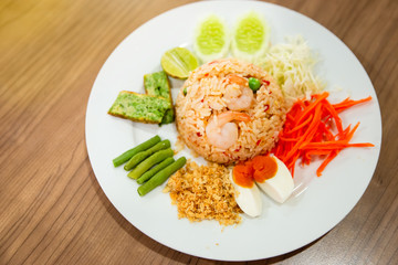Thai Spicy Shrimp Paste Fried Rice,Mixed vegetable such as carrot,cucumber,Yardlong beans, lime and boiled egg in white dish on wood table