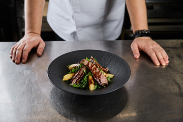 partial view of chef standing near dish with arugula, meat and potato at kitchen in restaurant