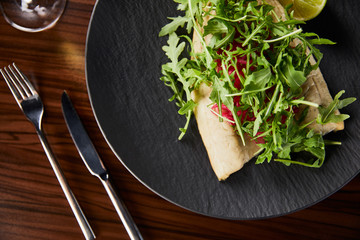 top view of tasty restaurant fish steak with lime and arugula on wooden table near cutlery