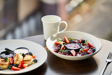 traditional Ukrainian varenyky and syrniki with berries served in white plate with sauce near cutlery and cup on wooden table in restaurant