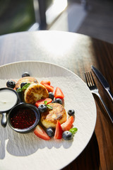 traditional Ukrainian dish syrniki with strawberry, blueberry served with jam, sour cream and cutlery on wooden table in sunlight