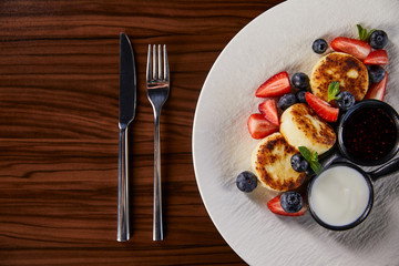 top view of traditional Ukrainian dish syrniki with strawberry, blueberry served with jam and sour cream on wooden table near cutlery