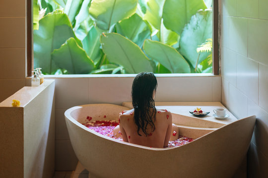 Back view of a young woman bathing in a health spa's flower bath full of petals. Luxury spa day with jungle view. Wellbeing, body care and beauty treatment concept
