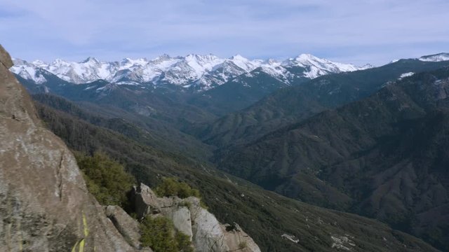 Drone shot panning around a massive granite wall and revealing the beautiful wilderness that is found in the Sierra-Nevada Mountains. You can see for miles of forests and snow-capped mountains.