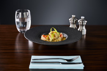 selective focus of delicious restaurant soup with shrimps in black plate served on napkin with cutlery and water on black background