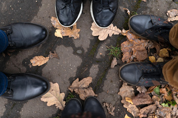 Four pairs of legs in boots and sneakers on the pavement against a background of yellow oak fallen leaves. Concept of autumn walk with friends. Togetherness. View from above