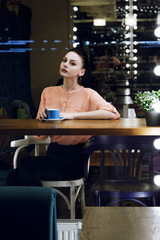 portrait of young woman drinking coffee at table in cafe through the window