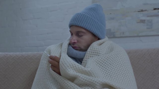 ill mean in warm hat wrapping in blanket and measuring temperature