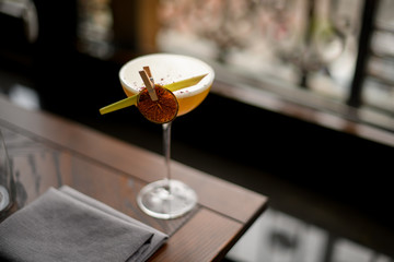 Yellow alcoholic cocktail in the glass with a dried lemon and tropical leaf in the clothespin on the table