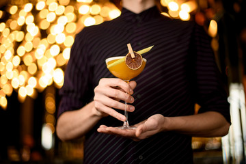 Male bartender in the purple shirt serving yellow alcoholic cocktail in the glass with a dried...