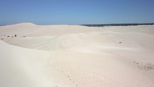 An aerial clip flying over a girl standing in the sand looking over the sand dune during mid-day.