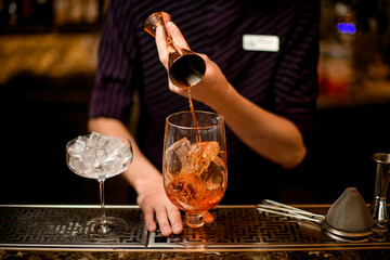 Bartender pouring a alcoholic drink from the jigger to a cocktail glass with ice cubes