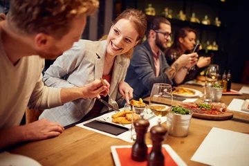 Foto op Plexiglas Knappe blanke gember die eten uit het bord van zijn vriendin haalt terwijl hij in het restaurant zit voor het avondeten. Op de achtergrond zijn hun vrienden. © chika_milan