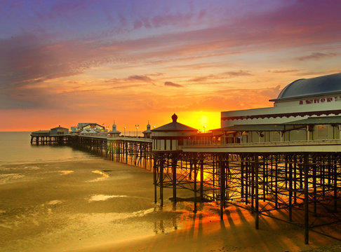 the sun setting over the historic north pier in blackpool with glowing golden light reflected on the beach and colourful twilight sky