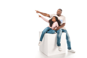 smiling african american father and daughter looking away and pointing with finger while sitting on cube together on white background