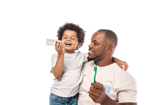 cheerful african american boy looking at toothpaste near happy father holding toothbrush isolated on white