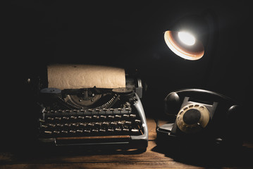 Old telephone and typewriter on the wooden desk in the light of the lamp background.