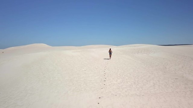 An aerial clip following and flying over a girl running in the sand revealing the sand dunes during mid-day.