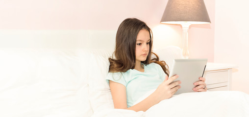 Obraz na płótnie Canvas Beautiful teenage girl looking and using tablet while lying in comfort bed in the morning in bedroom entertaining with mobile apps, portrait head shot