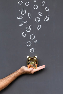Concept of saving money with a golden piggy bank held in a hand in front of a blackboard with hand drawn coins