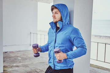 Young sportive man is doing exercises outside with dumbbells while posing for photographer.