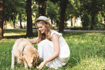 beautiful girl in white dress and straw hat petting golden retriever while sitting on meadow and looking at dog