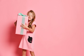 Fototapeta premium Christmas and New Year. Blonde kid girl in pink dress carrying a big long-awaited gifts box, hugging on pink. Banner
