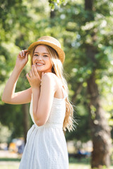 selective focus of beautiful young woman in white dress and straw hat smiling and looking at camera