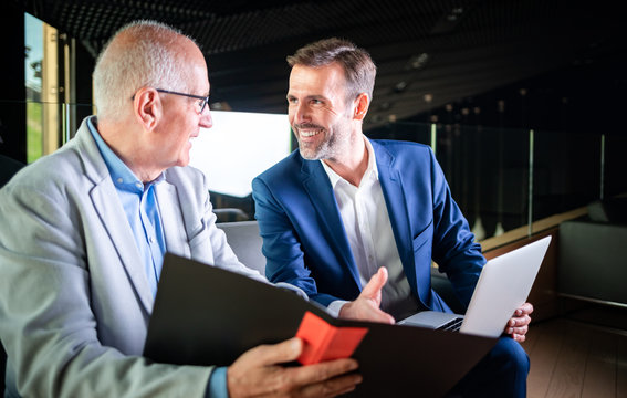 Younger And Older Businessman Talking About Documents During Meeting In Modern Business Lounge