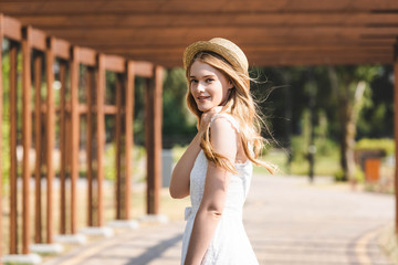 Obraz na płótnie Canvas selective focus of beautiful girl in white dress and straw hat walking near wooden construction and looking at camera
