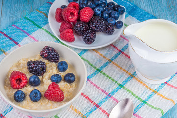 Healthy vegetarian breakfast - a bowl of oatmeal with berries, a plate with raspberries, blueberries, blackberries and a milk jug on a table with a napkin